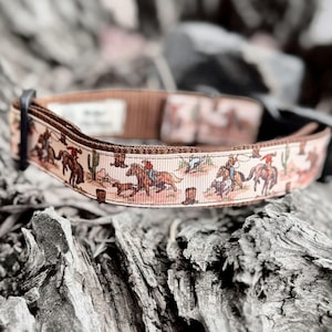 May include: A tan dog collar with a repeating western-themed pattern featuring cowboys, horses, cacti, and dogs. The collar has a brown backing and a black buckle. The design is printed on a ribbon-like material.