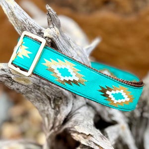 May include: A turquoise nylon collar featuring a repeating geometric pattern in white and metallic gold. The collar includes a rectangular gold-toned metal buckle and dark brown nylon webbing edges, providing a secure and adjustable fit.
