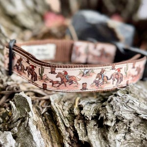 May include: A tan dog collar with a repeating western-themed pattern. The pattern features cowboys on horseback, cacti, and cowboy hats. The collar has a black buckle and a matching leash attachment.