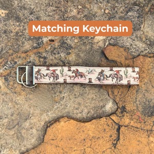 May include: A beige keychain with a repeating pattern of cowboys on horseback, cacti, and boots. The keychain has a silver metal clasp and the text "Matching Keychain" in an orange rectangle.