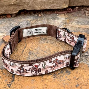 May include: Brown dog collar with a cowboy and desert landscape pattern. The collar has a black plastic buckle and a label that reads "Mabel Whoodle" with a dog illustration. The collar is designed for dogs.