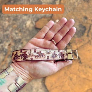 May include: A beige keychain wrist strap with a repeating pattern of cowboys on horseback, cacti, and other western-themed imagery. The strap has a gold-colored metal clasp and a matching keychain. Text on the image says "Matching Keychain."