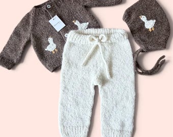 Puede incluir: Un conjunto de tres piezas de punto que incluye un cárdigan marrón de manga larga con bordado de pato blanco, pantalones color crema con cordón en la cintura y un gorro marrón a juego con bordado de pato blanco, todo con un patrón de punto.