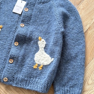 Könnte beinhalten: Eine blaue Strickjacke mit einer weißen, gestickten Ente mit gelben Füßen und Schnabel. Das Kleidungsstück hat einen gerippten Rundhalsausschnitt, lange Ärmel und eine Knopfleiste auf der Vorderseite mit hellbraunen Holzknöpfen. Ein weißes rechteckiges Etikett ist befestigt.