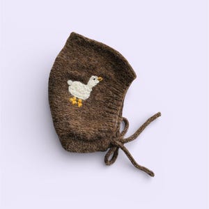 May include: A brown knit baby bonnet with a white embroidered duck design. The bonnet has a tie closure and is made of a soft, textured material. The duck has yellow feet and beak.