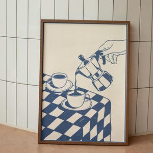 Może przedstawiać: Oprawiony wydruk z niebiesko-białą ilustracją wlewanego kawy. Grafika przedstawia dłoń wlewającą kawę z kawiarki na stół z niebieskim, w kratę obrusem. Na stole znajdują się również dwie filiżanki i spodki.