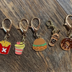 May include: Five gold-colored keychains with different food charms. The charms are a red and yellow french fry box, a pink and yellow cupcake, a green and brown hamburger, a brown iced coffee, and a brown donut.