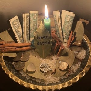 Pode incluir: Uma vela verde acesa está em uma tigela decorativa com notas, moedas, folhas de louro, paus de canela e cristais. Uma folha com o texto "lunar astrid" está no centro. A tigela é preenchida com arroz.