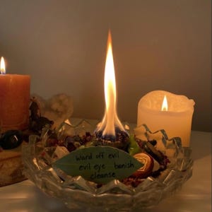 May include: A lit candle with a long, bright flame sits in a clear glass bowl filled with herbs and crystals. A handwritten note on a leaf reads "Ward off evil, evil eye, banish, cleanse." Two other lit candles are in the background.