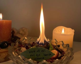 May include: A lit candle with a long, bright flame sits in a clear glass bowl filled with herbs and crystals. A handwritten note on a leaf reads "Ward off evil, evil eye, banish, cleanse." Two other lit candles are in the background.