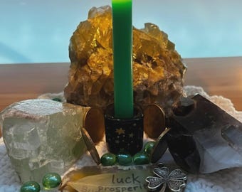 May include: A lit green candle in a black holder with gold stars, surrounded by crystals, coins, and green glass beads. A leaf-shaped stone with the words "luck, prosperity, business, wealth" and a four-leaf clover charm are also present.