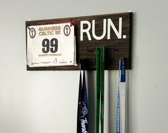 Könnte beinhalten: Ein Holzschild mit dem Wort "RUN." in weißen Buchstaben. Das Schild hat Haken zum Aufhängen von Medaillen und einer Startnummer. Drei Medaillen hängen an den Haken, eine ist blau und weiß, eine ist grün und eine ist golden. Die Startnummer ist weiß mit dem Text "GUINNESS CELTIC 5K" und der Nummer "99".