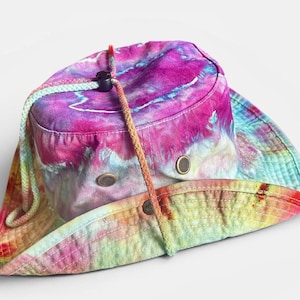May include: A tie-dye bucket hat featuring a vibrant blend of magenta, purple, blue, and yellow colors. The hat includes a braided chin strap with a black plastic toggle and two metal ventilation eyelets on each side of the crown.