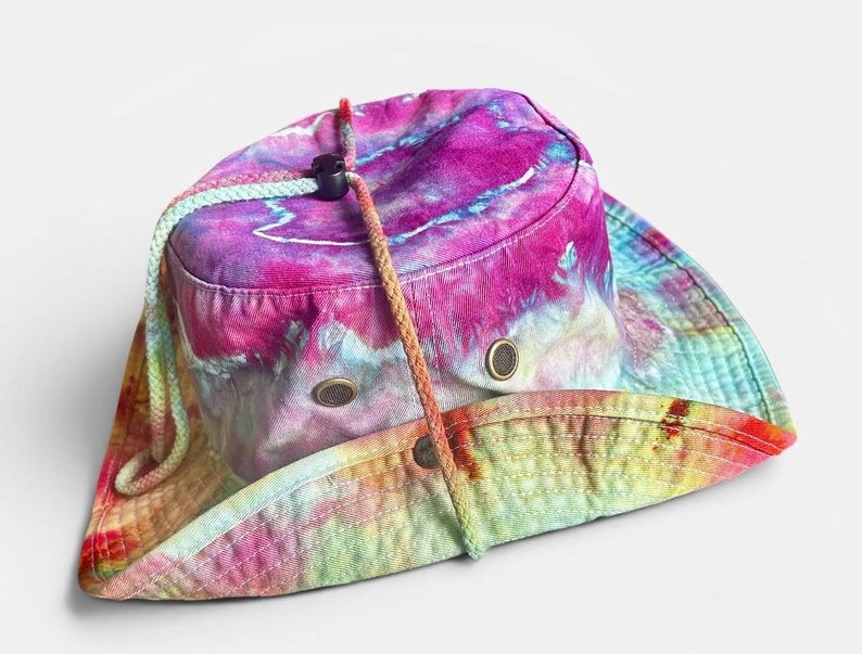 May include: A tie-dye bucket hat featuring a vibrant blend of magenta, purple, blue, and yellow colors. The hat includes a braided chin strap with a black plastic toggle and two metal ventilation eyelets on each side of the crown.
