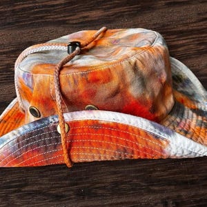 May include: A wide-brimmed bucket hat featuring a vibrant tie-dye pattern in shades of orange, red, and grey. The hat includes a braided brown chin strap with a black plastic toggle and metal ventilation eyelets on the side of the crown.