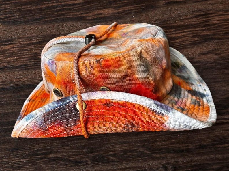 May include: A wide-brimmed bucket hat featuring a vibrant tie-dye pattern in shades of orange, red, and grey. The hat includes a braided brown chin strap with a black plastic toggle and metal ventilation eyelets on the side of the crown.