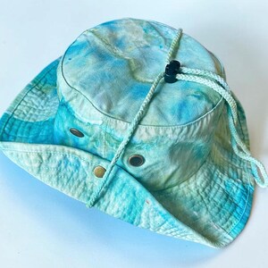 May include: A tie-dye bucket hat in shades of light blue, turquoise, and white. The hat features a wide brim, a braided chin strap with a black plastic toggle, and two metal ventilation eyelets on each side of the crown for airflow.
