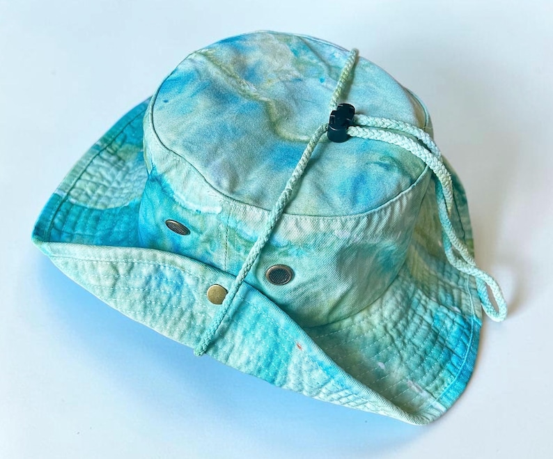 May include: A tie-dye bucket hat in shades of light blue, turquoise, and white. The hat features a wide brim, a braided chin strap with a black plastic toggle, and two metal ventilation eyelets on each side of the crown for airflow.