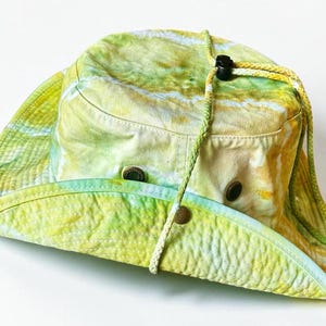 May include: A wide-brimmed bucket hat featuring a tie-dye pattern in shades of yellow, lime green, and white. The hat includes a braided chin cord with a black plastic toggle, metal ventilation eyelets on the sides, and a snap button on the brim.