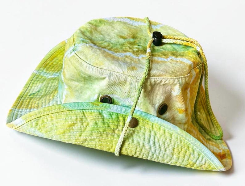 May include: A wide-brimmed bucket hat featuring a tie-dye pattern in shades of yellow, lime green, and white. The hat includes a braided chin cord with a black plastic toggle, metal ventilation eyelets on the sides, and a snap button on the brim.