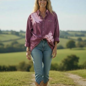 May include: A woman wearing a long-sleeved, button-down, red plaid shirt with white accents, light blue jeans, and brown cowboy boots. She is walking on a dirt path in a field with green hills in the background.