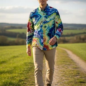 May include: A button-down shirt with a vibrant tie-dye pattern in shades of blue, yellow, and red. The shirt is paired with khaki pants and brown hiking boots. The person is walking on a dirt path through a green field.