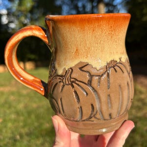 Peut inclure: Une tasse en céramique avec un motif de citrouille. La tasse présente une base marron avec des citrouilles sculptées, passant à un dégradé beige et orange en haut. La poignée est orange avec des accents marron. La tasse est tenue dans une main.