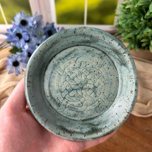 Può includere: Un piccolo piatto rotondo in ceramica con smalto turchese e un disegno a nodo celtico al centro. Il piatto ha una texture maculata e un bordo leggermente rialzato. Lo sfondo include una finestra, fiori blu e vegetazione.