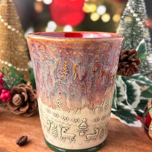 May include: A handcrafted ceramic coffee mug with a festive Christmas theme. The mug features a textured design with deer, trees, and snowflakes. The mug has a green base, a red interior, and is surrounded by coffee beans.