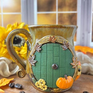 May include: A ceramic mug with a glossy brown glaze featuring a sculpted green circular door on the front. The door is framed by a textured wood-like border adorned with small brown leaves, yellow flowers, and a bright orange pumpkin at the base.
