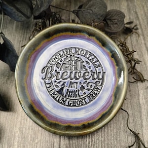 Könnte beinhalten: Ein Keramikteller mit erhöhtem Rand und glänzendem Finish. Der Teller zeigt den Text "Foolish Mortals Brewery" in einem kreisförmigen Design, mit einer Gebäudeillustration und einer Fledermausgrafik. Der Rand hat einen Farbverlauf aus Gold, Lila und Grün.