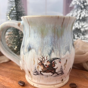 Op de afbeelding: Een keramische koffiemok met een winterse scène met een kabouter die op een rendier rijdt. De mok heeft een crèmekleurige basis met blauwe en groene glazuurdruppels. Koffiebonen liggen verspreid op het houten oppervlak.