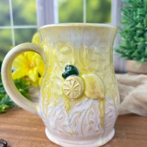 Puede incluir: Una taza de cerámica con un esmalte amarillo y blanco, con diseños de limones en relieve. La taza tiene un asa curva y está decorada con una hoja verde y una rodaja de limón. Granos de café están esparcidos sobre la superficie de madera.