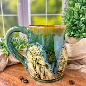 Puede incluir: Taza de cerámica hecha a mano con un esmalte verde y dorado. La taza presenta un diseño en relieve de setas y follaje. El asa es verde. Varios granos de café están esparcidos alrededor de la taza sobre una superficie de madera.