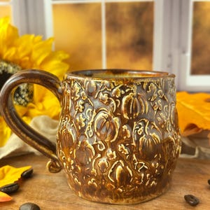 May include: A brown ceramic mug featuring a raised, textured relief pattern of pumpkins and leaves across its entire exterior surface. The mug has a glossy finish with darker brown accents in the recessed areas and a curved handle on the side.