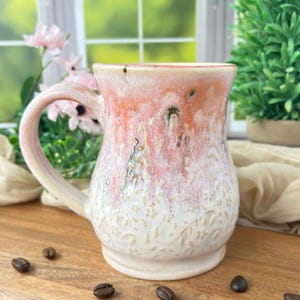 May include: A handmade ceramic mug with a peach and white glaze. The mug features a large handle and a floral pattern embossed on the lower half. Coffee beans are scattered around the base of the mug.