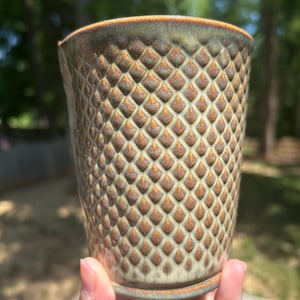 Op de afbeelding: Een handgemaakte keramische beker met een textuur van diamantpatroon. De beker heeft een lichtgroene basis met bruine en gouden accenten. De rand en de basis hebben een donkerdere bruine glazuur. De beker wordt in de hand van een persoon gehouden.