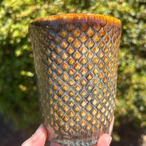 May include: A ceramic tumbler featuring a textured, raised diamond pattern across its surface. The glaze transitions from a deep, warm amber at the rim to a muted, earthy green and brown finish toward the base, creating a multi-tonal appearance.