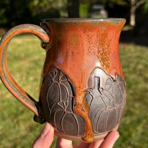 May include: A ceramic mug featuring a dark brown base with carved pumpkin designs and a glossy, dripping orange and amber glaze that covers the upper portion and handle, creating a distinct contrast between the textured and smooth surfaces.
