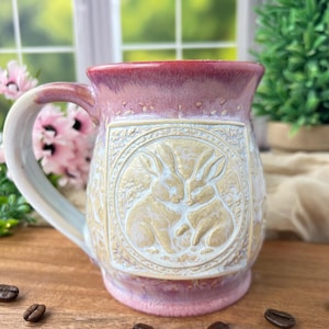 May include: A handmade ceramic mug with a pink and cream glaze. The mug features a raised relief design of two rabbits within a circular frame. The handle is cream-colored, and coffee beans are scattered around the base.