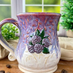 Puede incluir: Una taza de cerámica con esmalte púrpura y crema, que presenta un relieve tridimensional de moras de color púrpura oscuro y hojas verdes en la parte frontal. La taza tiene un asa curva y un borde ancho con acabado brillante.