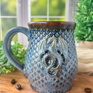 May include: A ceramic mug featuring a blue and brown gradient glaze with a textured dragon scale pattern across the exterior. A sculpted, raised dragon figure with wings is centered on the front. The mug has a wide handle and a dark brown rim.