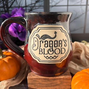 Pode incluir: Uma caneca de cerâmica com esmalte vermelho escuro e castanho, com um rótulo creme com as palavras "Dragon's Blood" e uma ilustração de um dragão. A caneca está colocada num bloco de madeira com abóboras e grãos de café por perto.