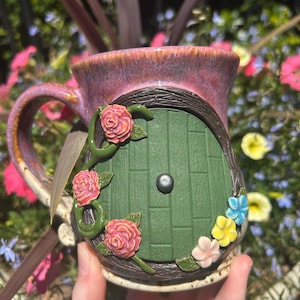 Peut inclure: Une tasse en céramique avec une finition émaillée violette et rose, ornée d'une porte verte sculptée avec un bouton central. La porte est encadrée par des textures en bois brun foncé et décorée de trois roses roses et de petites fleurs bleues, jaunes et blanches.