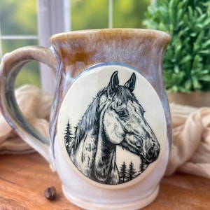 Könnte beinhalten: Eine Keramik-Kaffeetasse mit einem Pferdeporträt. Die Tasse hat eine braun-blaue glasierte Außenseite und eine weiße Innenseite. Auf der Vorderseite befindet sich eine Schwarz-Weiß-Illustration eines Pferdekopfes. Kaffeebohnen sind um die Tasse verstreut.