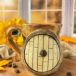 May include: A ceramic mug featuring a sculpted, light-colored wooden door design on the front. The mug has a glossy brown and tan glaze, a curved handle, and a circular door with vertical plank detailing and a small, dark central knob.