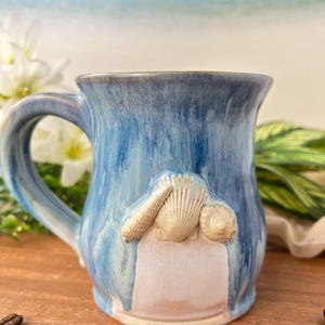 Puede incluir: Una taza de cerámica blanca con asa negra y el texto "But First, Coffee" en negrita negra. La taza se encuentra sobre un fondo borroso, resaltando su diseño limpio.
