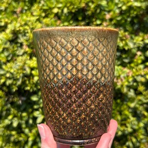 Op de afbeelding: Een cilindervormige keramische beker met een gestructureerd ruitpatroon over het oppervlak. Het glazuur verloopt van een licht olijfgroene kleur bij de rand naar een diep donkerbruin bij de basis, wat een verloopseffect met glans geeft.