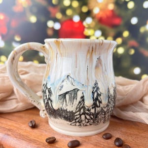 May include: Cream-colored ceramic mug with a mountain and forest design. The mug features a detailed illustration of a mountain range and pine trees in black and white. The handle is a light beige color, and coffee beans are scattered around the mug.