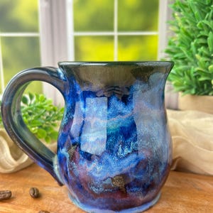 May include: A ceramic mug featuring a glossy glaze with swirling patterns of deep blue, violet, and black. The mug has a wide, curved handle and a rounded body, with a smooth, reflective surface that transitions between dark and light blue tones.